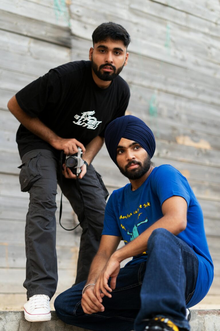 Two men, one with a camera, pose casually against a wooden wall, showcasing urban style.